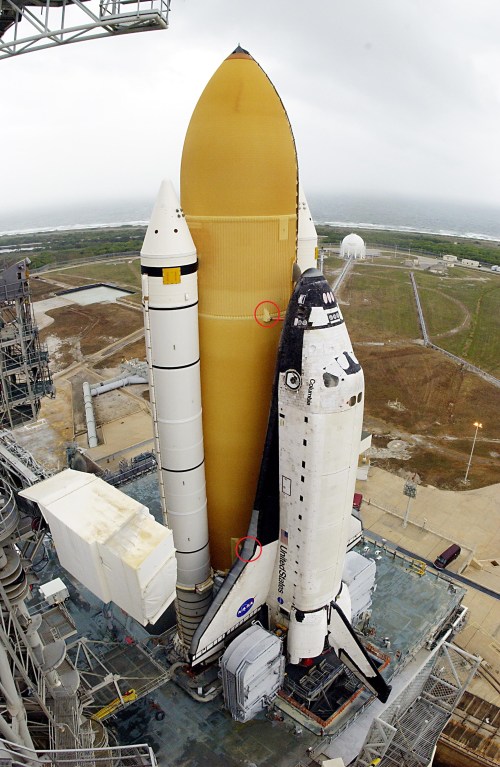 Space shuttle on launch pad, with external fuel tank and solid rocket boosters, ready for liftoff against a cloudy sky.