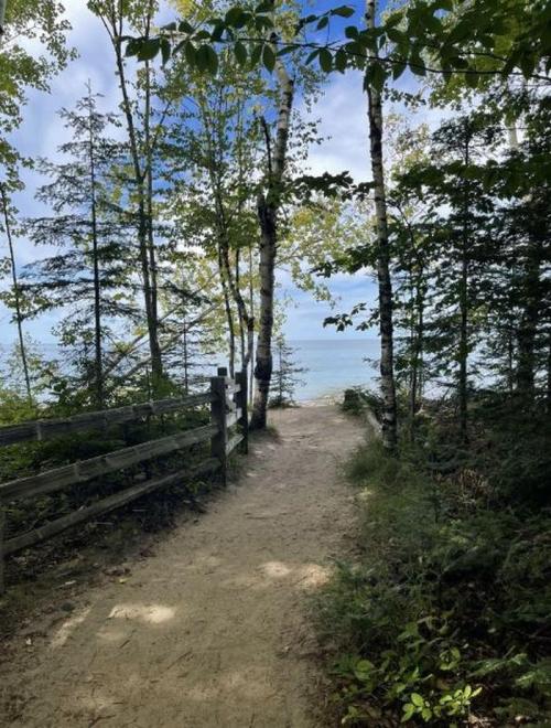 A sandy path through trees leads to a serene lakeside view under a partly cloudy sky.
