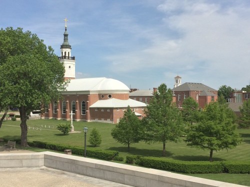 A view of a church with a tall steeple, surrounded by green trees and manicured lawns under a blue sky.