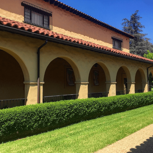 A sunny courtyard with arched columns, a manicured hedge, and a building featuring a tiled roof and windows.