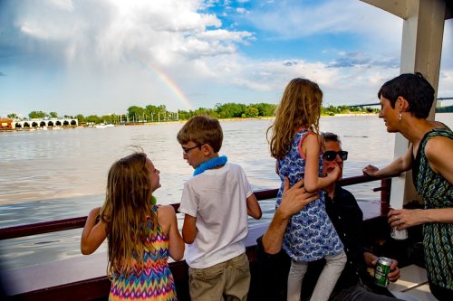 A family enjoys a boat ride by the water, with children playing and a rainbow visible in the background.