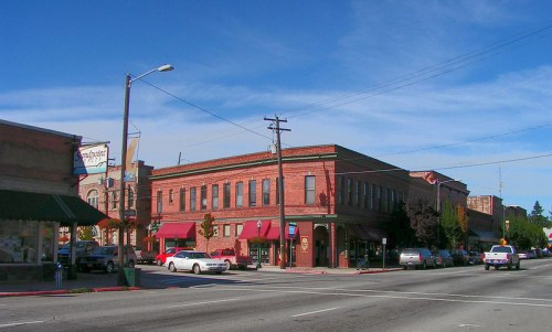 Historic brick buildings line a street under a clear blue sky, with parked cars and storefronts visible.
