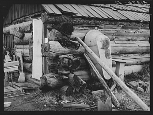 A rustic log cabin with a wooden door, tools, and debris scattered around the entrance.