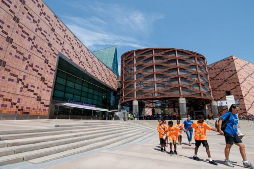 A modern building with a unique design, featuring children in orange shirts walking towards it on a sunny day.