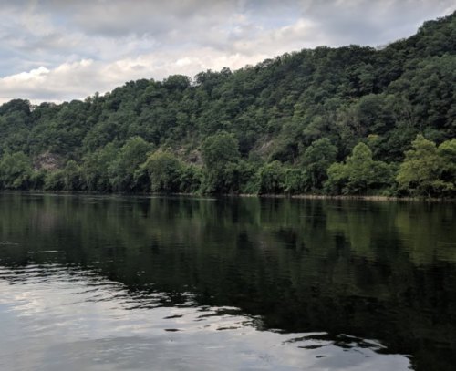 A calm river reflects lush green trees and a cloudy sky, surrounded by a wooded hillside.