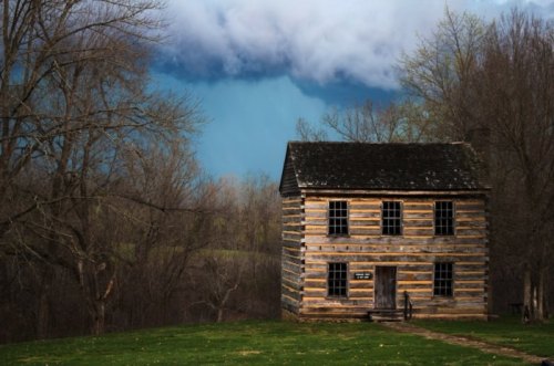 A rustic wooden house stands in a grassy area, with dark clouds looming in the background and trees surrounding the scene.