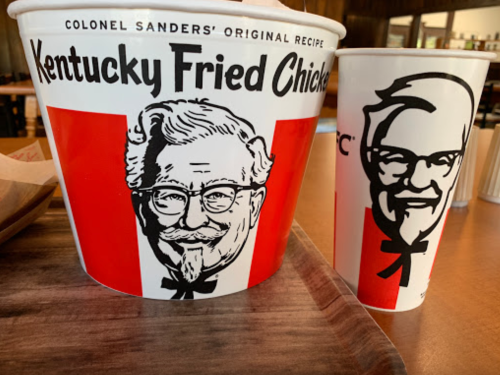 A bucket of Kentucky Fried Chicken and a drink cup featuring the Colonel Sanders logo.
