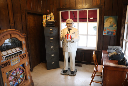A life-sized statue of a man in a white suit holding a bucket, inside a rustic room with wooden walls and vintage decor.