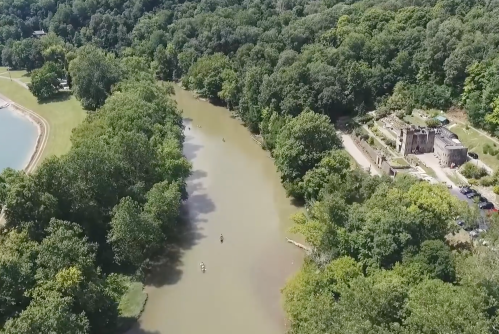 Aerial view of a river surrounded by lush greenery, with a historic building and a small beach nearby.