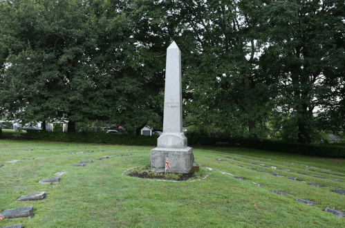 A tall stone obelisk stands in a grassy cemetery, surrounded by trees and grave markers, with a small American flag nearby.