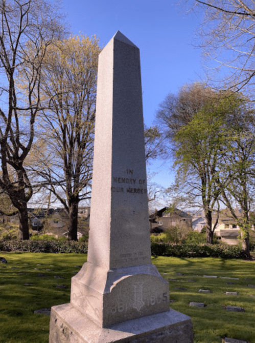 A tall stone monument in a cemetery, surrounded by trees, with the inscription "In Memory of Our Heroes."