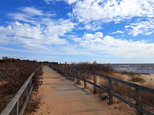 A wooden boardwalk leads through sandy terrain to a beach under a blue sky with scattered clouds.