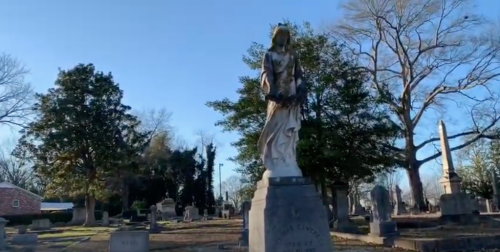 A tall statue of a woman in a cemetery, surrounded by trees and gravestones under a clear blue sky.