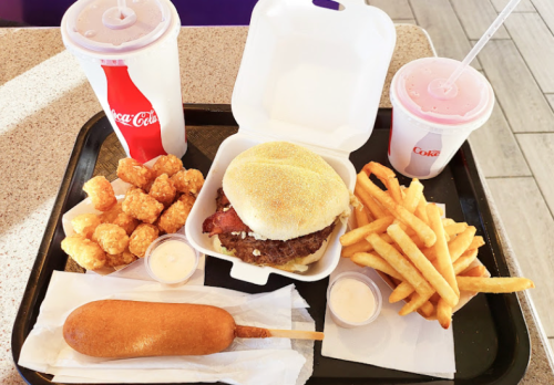 A tray with a burger, fries, tater tots, a corn dog, and two soft drinks.