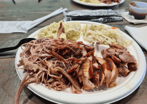 A plate of pulled pork, grilled chicken, coleslaw, and mashed potatoes on a wooden table.