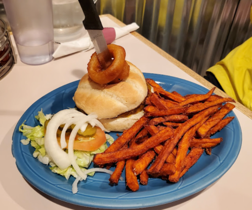 A plate with a sandwich topped with an onion ring, served with sweet potato fries and a side salad.