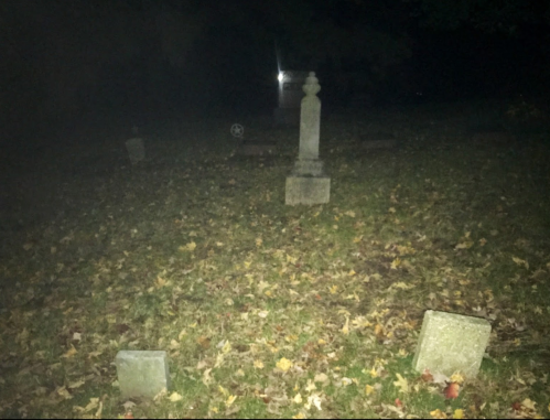 A dimly lit graveyard with scattered tombstones surrounded by fallen leaves at night.
