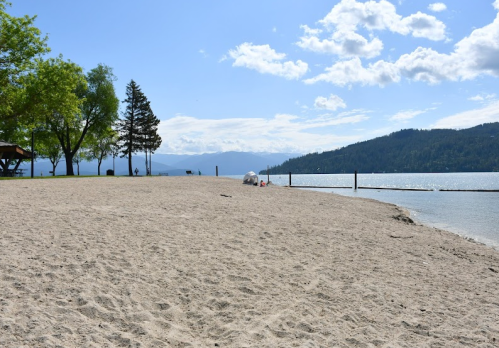 A sandy beach with trees, a calm lake, and distant mountains under a partly cloudy sky.