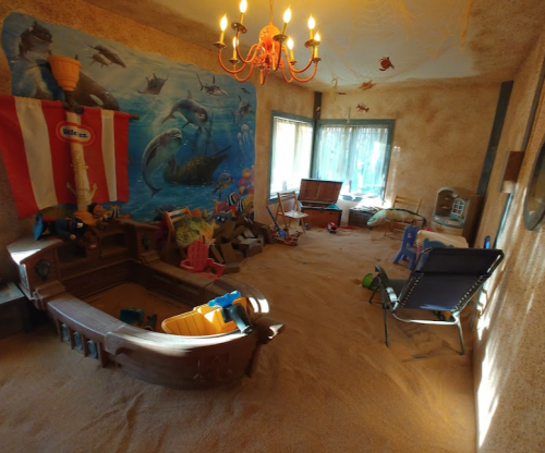 A playful room with sandy flooring, a boat, toys, and a mural of dolphins on the wall, illuminated by a chandelier.