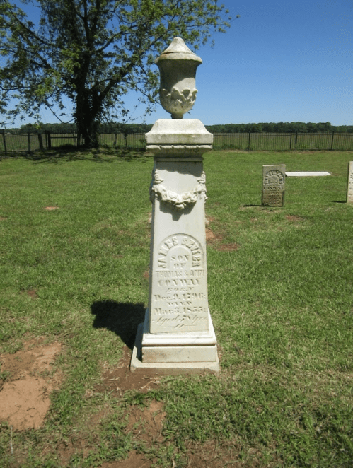 A white gravestone with inscriptions, surrounded by grass and trees under a clear blue sky.
