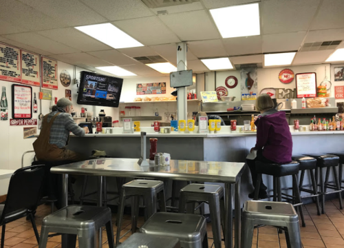 Two patrons sit at a diner counter, watching a TV, surrounded by vintage decor and condiments on the tables.