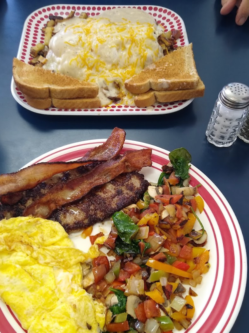 A plate with an omelet, bacon, and sautéed vegetables, alongside a dish of toast and gravy-covered potatoes.