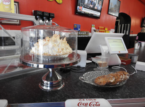 A glass display case with a meringue pie and a plate of cookies on a counter in a diner.
