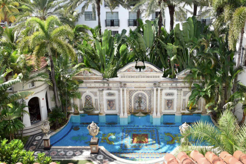 A lush tropical courtyard featuring a decorative pool surrounded by palm trees and ornate architecture.