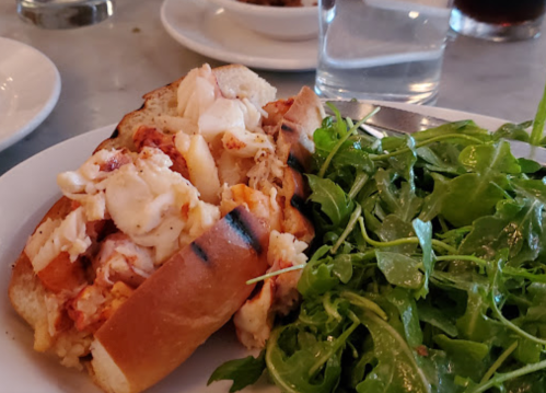 A lobster roll on a plate with a side of fresh arugula salad and a glass of water in the background.