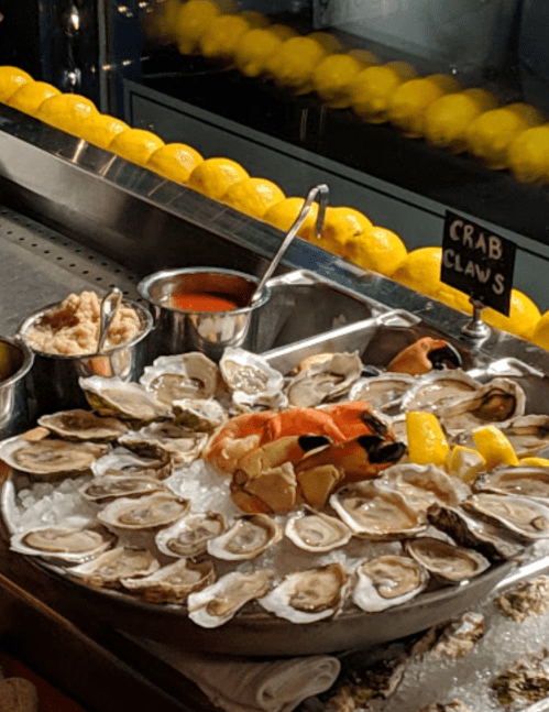 A seafood platter featuring oysters, crab claws, lemon, and dipping sauces on ice.
