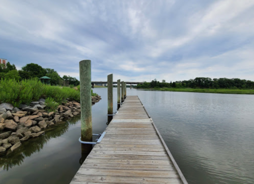 A wooden dock extends into a calm river, surrounded by greenery and under a cloudy sky.