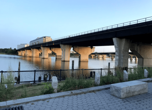A bridge spans over a calm river, with concrete pillars and a grassy bank in the foreground under a clear sky.