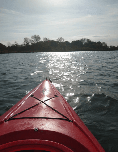 A red kayak on calm water, reflecting sunlight, with trees in the background under a clear sky.
