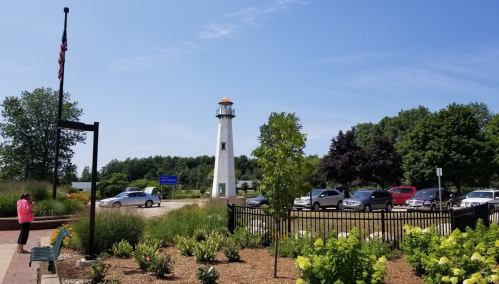 A lighthouse stands in a park with greenery, cars parked nearby, and a person in a pink jacket by the flagpole.