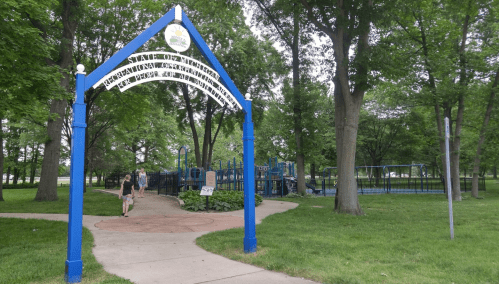 A blue archway leads to a playground surrounded by lush green trees and grass in a park.