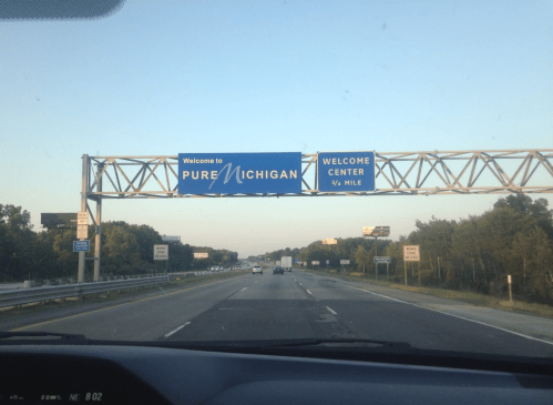 Sign reading "Welcome to Pure Michigan" with directions to a welcome center, viewed from a vehicle on the highway.