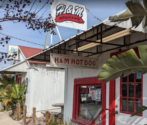 Exterior of H&M Hotdog Stand with a sign, featuring a red and white color scheme and tropical plants nearby.