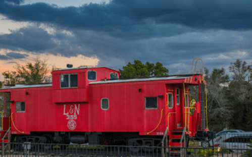 A red train caboose labeled "L&N 3155" sits against a cloudy sky, surrounded by trees.