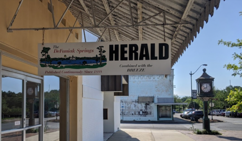 Sign for "The Defuniak Springs Herald," with a clock and buildings in the background. Published continuously since 1885.