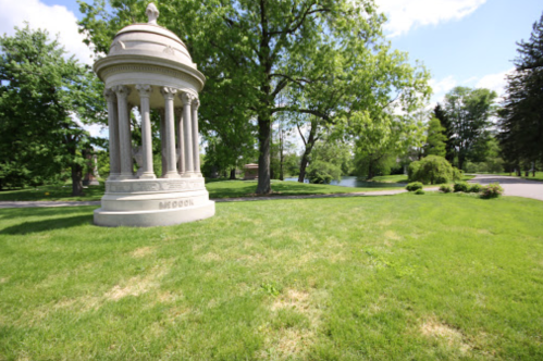 A stone gazebo surrounded by lush green grass and trees, with a pathway and water visible in the background.
