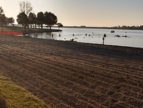 A sandy beach at sunset with people swimming and kayaking in the calm water, surrounded by trees.
