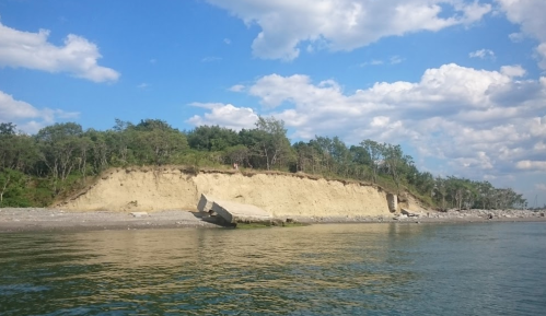 A sandy cliff with trees on top, reflecting in calm water under a blue sky with scattered clouds.