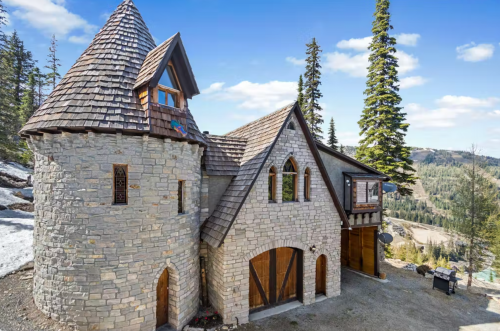 A charming stone house with a turret, surrounded by trees and mountains under a clear blue sky.