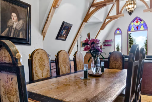 A rustic dining room with a wooden table, ornate chairs, and a vintage portrait on the wall, featuring stained glass windows.
