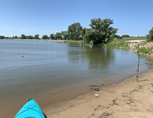 A calm lake scene with a kayak in the foreground, sandy shore, and lush greenery along the water's edge.