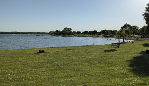 A serene lakeside view with green grass, trees, and people enjoying the water on a sunny day.