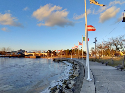 A scenic waterfront view with flags from various countries, ice on the water, and a clear blue sky.