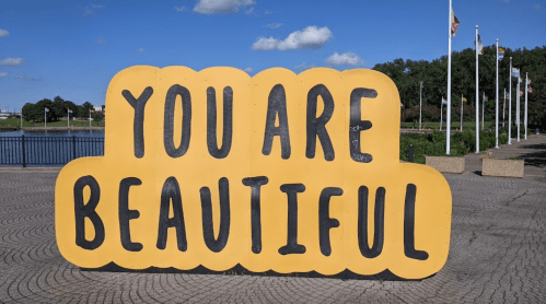 A large, bright yellow sign with bold black letters reads "YOU ARE BEAUTIFUL" against a blue sky and park backdrop.