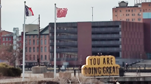 A colorful sign reading "YOU ARE DOING GREAT" in front of buildings and flags near a waterfront.