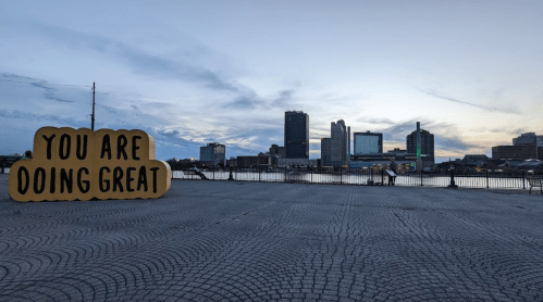 A large sign reading "YOU ARE DOING GREAT" in front of a city skyline at sunset by the water.